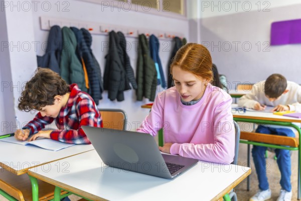 Elementary school students studying at individual desks, girl using laptop, boy writing in notebook, winter coats hanging on wall