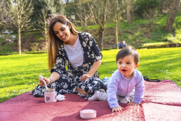 Happy mother preparing a meal from a thermos for her baby during a picnic on a blanket in a sunny park