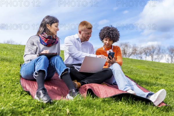 Three students collaborating on a project, using laptop, tablet, and smartphone, enjoying sunny weather in a park