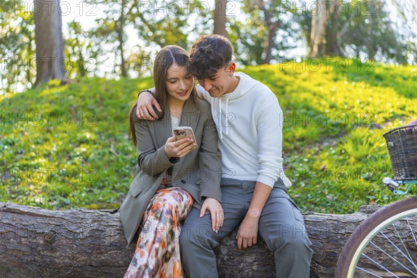 Smiling young couple sitting on a log in a park, looking at a smartphone together, surrounded by lush greenery and a bicycle nearby