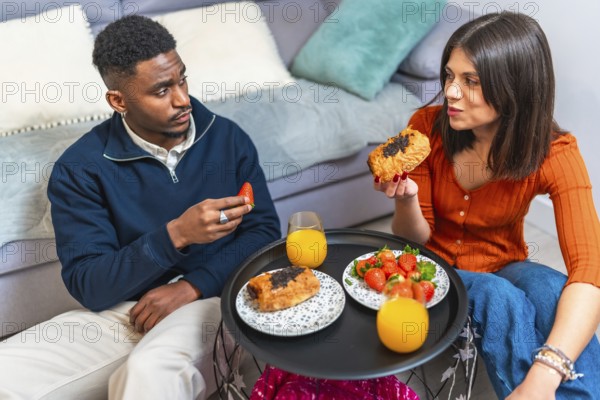 Young multiethnic couple eating pastries and strawberries with orange juice, enjoying breakfast at home
