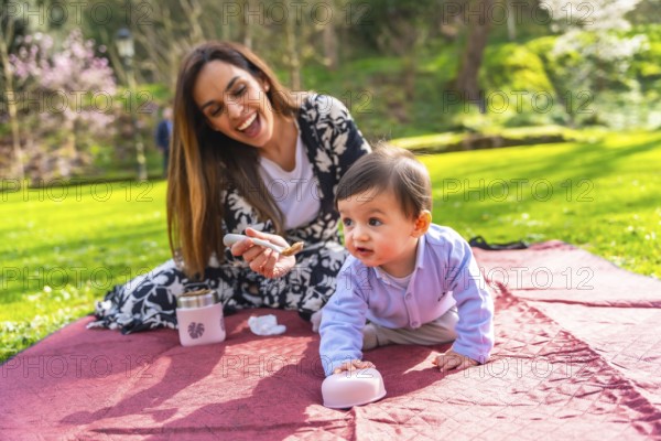 Happy mother feeding her baby solid food with a spoon, sitting on a blanket during a picnic in a park