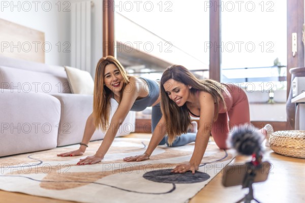 Two smiling women practicing yoga together in their living room, recording an online class with a smartphone