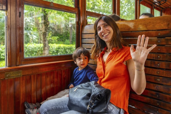 A woman and a child are sitting on a train. The woman is wearing an orange shirt and is waving at the camera