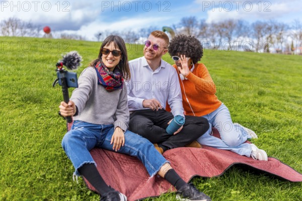 Three content creators are sitting on blanket in park, filming video blog using smartphone with external microphone, having fun and enjoying sunny day
