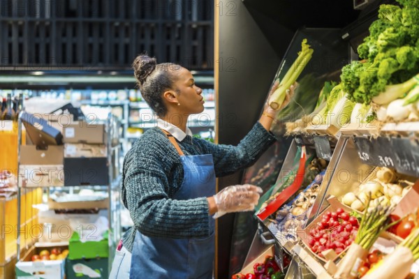 Supermarket employee wearing gloves arranging organic celery on a shelf, surrounded by various fresh produce in a grocery store