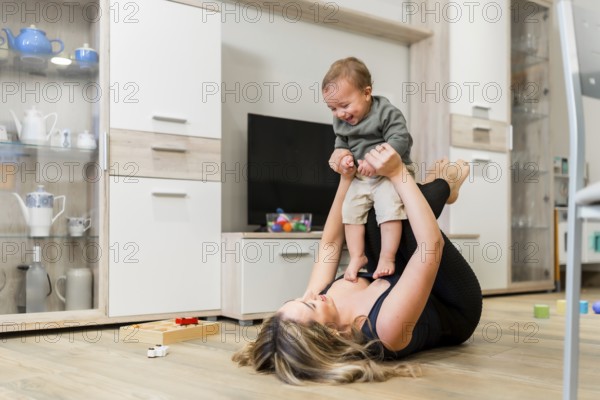 Young mother lying on the floor lifting her baby in the air while playing together in the living room at home