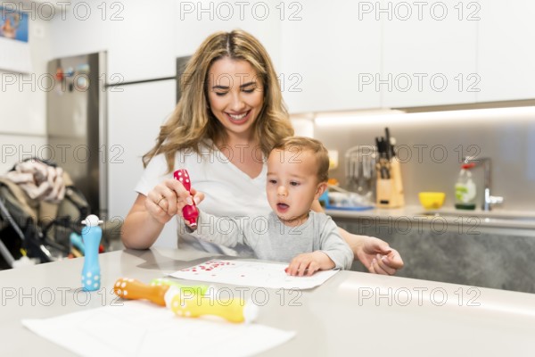 Smiling mother and baby boy having fun drawing together with colorful dot markers in a modern kitchen