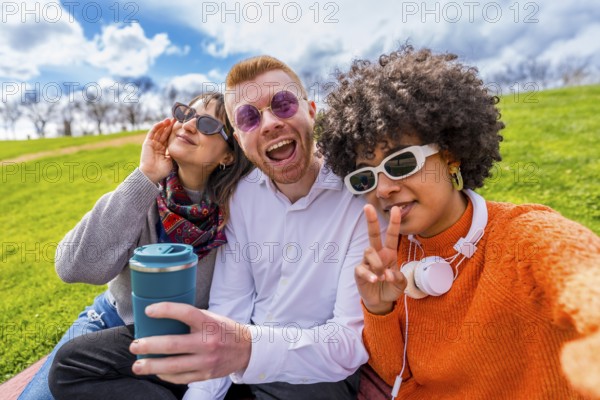 Three cheerful multi ethnic friends making funny faces and gesturing while taking a selfie with reusable coffee cup in a city park