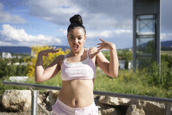 A woman is posing for a picture with her arms raised and a ponytail. She is wearing a pink top and shorts. Scene is energetic and confident