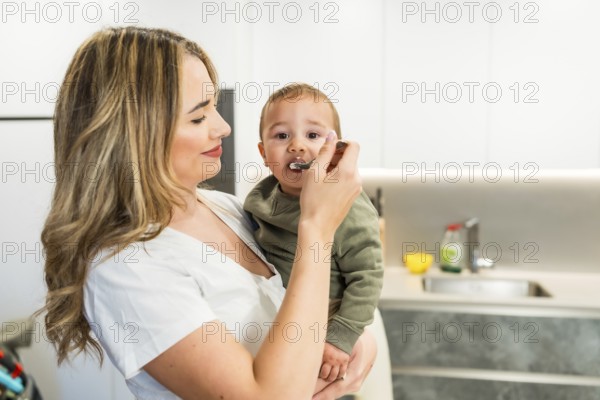 Loving mother feeding her baby son with a spoon in a bright modern kitchen, enjoying a tender moment of family bonding
