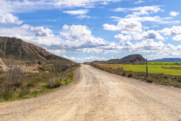 Scenic view of a gravel road stretching through the bardenas reales desert landscape under a bright blue sky with fluffy white clouds