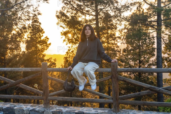 Young woman sits on a wooden fence, taking in the breathtaking sunset over the forested landscape of moya, gran canaria