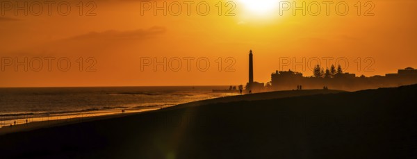 Tourists enjoying a beautiful sunset over maspalomas dunes in gran canaria, spain, with the iconic lighthouse silhouetted against the orange sky