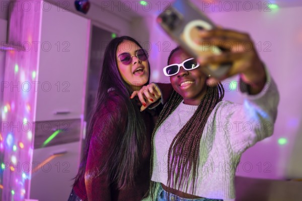 Two multi ethnic female friends wearing sunglasses and enjoying a home party, taking selfies with smartphone under colorful disco lights