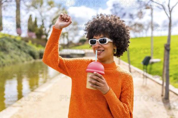 Happy young woman with curly hair and orange sweater, enjoying a smoothie in a park by the river, raising her arm in a carefree gesture
