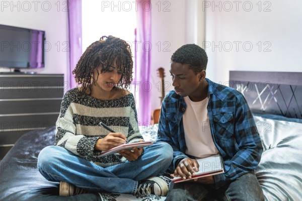 Two focused college students are sitting on a bed, sharing notes and preparing for exams in their dorm room