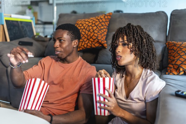 Relaxed couple enjoying a movie night at home, sitting on the floor in front of their sofa and eating popcorn from striped buckets