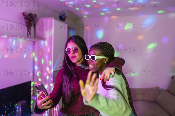 Two teenage girls wearing sunglasses are embracing and taking a selfie with smartphone at a home party with colorful disco lights