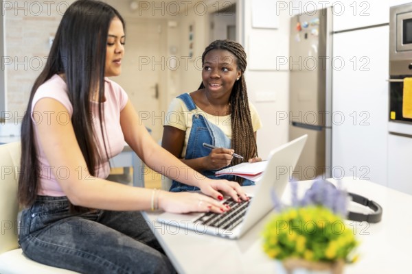 Two teenage girls collaborating on homework, using a laptop and taking notes in a modern kitchen