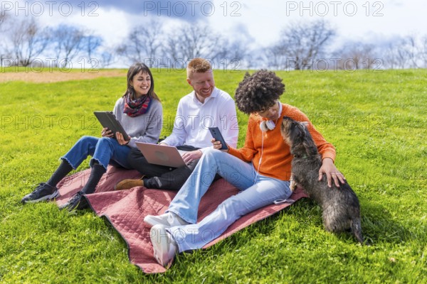 Three young coworkers are taking a break from work outdoors, using laptop, tablet and smartphone, while petting a dog in a park