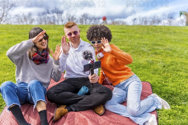Three content creators waving cheerfully at the camera while filming a video blog outdoors in a sunny park, enjoying their time together