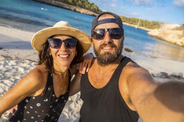 A man and woman are posing for a picture on a beach. The man is wearing a black tank top and sunglasses, while the woman is wearing a straw hat and sunglasses. They both have smiles on their faces
