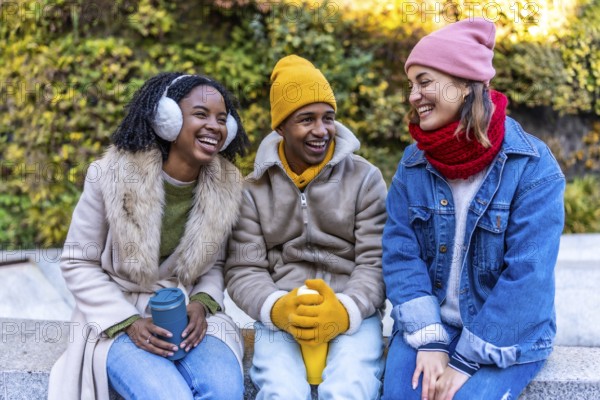 Three cheerful young multi ethnic friends sitting outdoors in the city on a cold winter day, enjoying reusable coffee cups