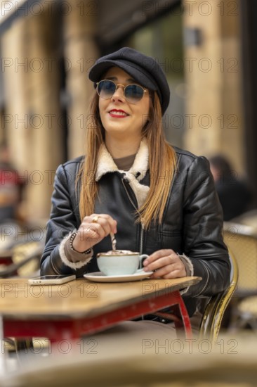 Young woman wearing sunglasses and hat stirring cappuccino at a parisian outdoor cafe, enjoying a relaxing moment