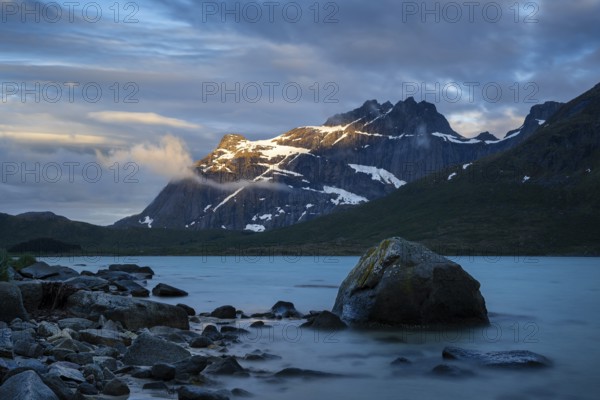 Landscape with sea and mountains on the Lofoten Islands, view over the fjord Flakstadpollen to mountains. Rocks in the foreground. At night at the time of the midnight sun, some clouds in the sky, the sun shining on the mountain. Long exposure. Early summer. Flakstadoya, Lofoten, Norway, Europe