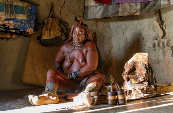 Traditional Himba woman applying ochre paint for body colouring, interior of the first woman's mud hut in a Himba village, traditional containers for ochre paint and woven frame with leather for body care with smoke, near Opuwo, Kaokoveld, Kunene, Namibia, Africa