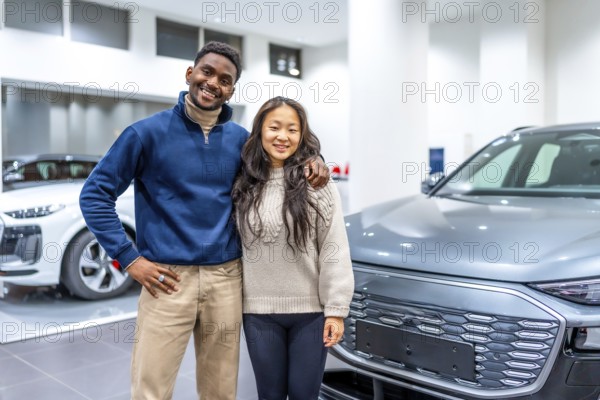 Smiling multiethnic couple buying a new electric vehicle at car dealership, embracing each other next to their new electric car