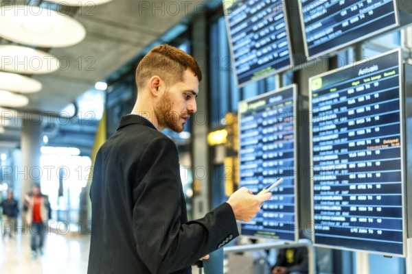 Businessman checking flight information on an airport display while using a smartphone, staying connected during travel and planning his journey