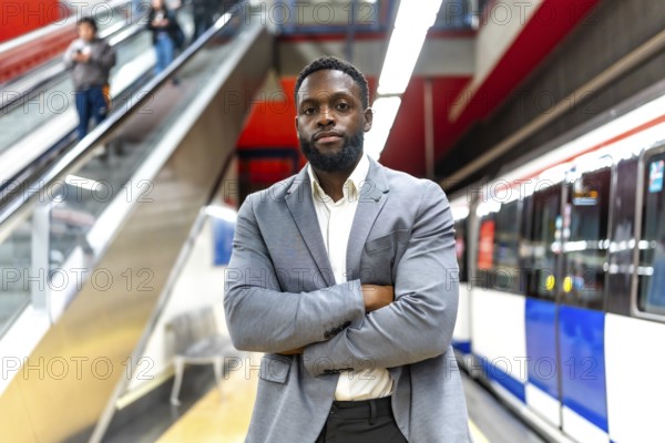 Young black businessman standing with crossed arms in subway station, waiting for transportation, commuting to work