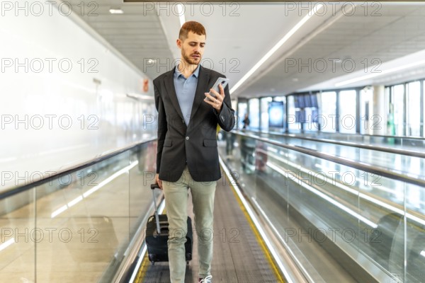 Businessman pulling suitcase and using smartphone while walking on moving walkway at airport