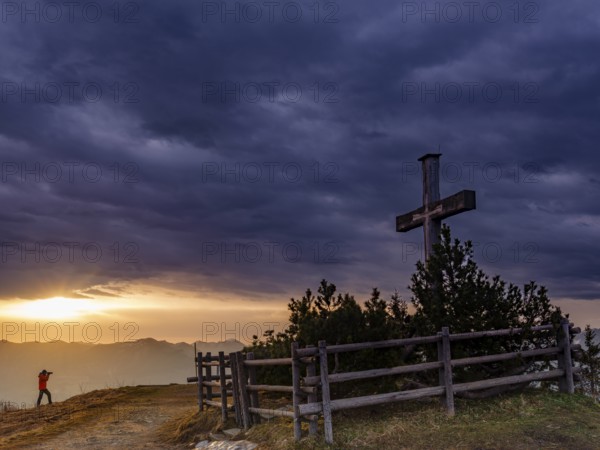 Photographer next to the summit cross on the Roßfeld in a dramatically overcast sky, the sun shines through a gap behind, Roßfeldpanoramastraße, Oberau, Berchtesgadener Land, Upper Bavaria, Bavaria, Germany, Europe