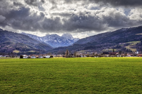 Dramatic Alpine landscape with green meadow in the foreground and cloudy sky over a village, Bad Feilnbach