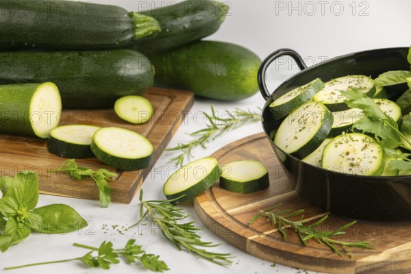 Sliced and seasoned courgettes with fresh herbs in a black pot, next to stacked courgettes on a wooden board