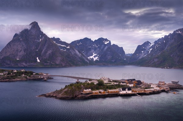 The village of Sakrisoy with its typical yellow or ochre-coloured wooden houses on wooden stilts (rorbuer) . A bridge over the sea. Mount Olstinden in the background on the left. At night at the time of the midnight sun. Clouds in the sky. Early summer. Sakrisoy, Moskenesoya, Lofoten, Norway, Europe