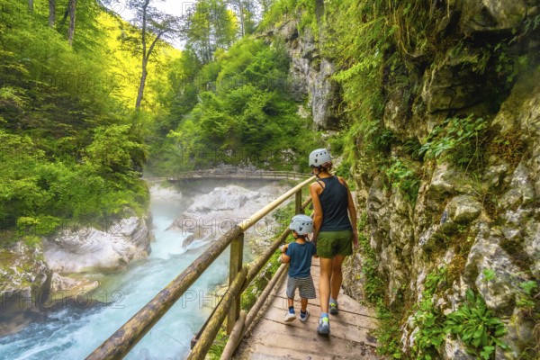 Mother and son wearing helmets walking on wooden path and enjoying breathtaking view of radovna river flowing through vintgar gorge in slovenia
