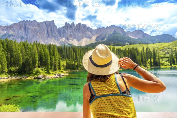 Woman wearing straw hat admiring lake carezza with latemar mountain range in background, dolomites, italy