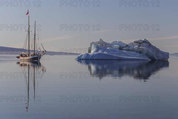 Traditional sailing ship reflected in fjord next to iceberg, sunny, Ittoqqortoormiit, Scoresby Sund, East Greenland, Greenland, North America