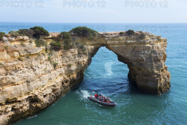 A boat with tourists sails through a natural rock arch over the clear blue sea, Arco de Albandeira, Lagoa, Algarve, Portugal, Europe