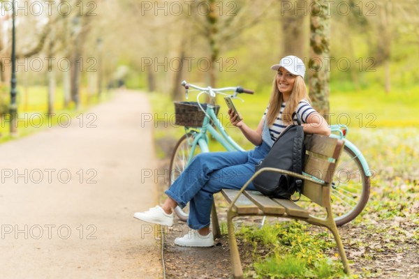Happy tourist is using her smartphone while resting on a bench in a park with her bicycle next to her