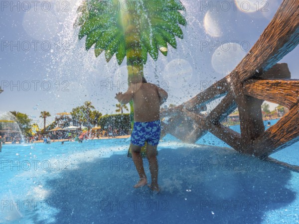 Child enjoying refreshing splashes under a decorative palm tree in a water park, creating joyful summer memories