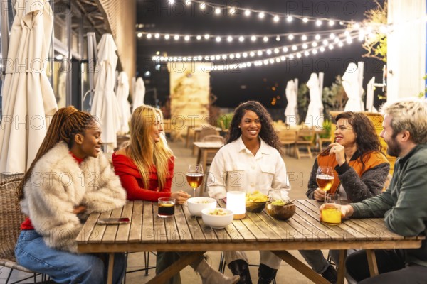 Multi-ethnic friends sitting on a bar terrace drinking celebrating together at night