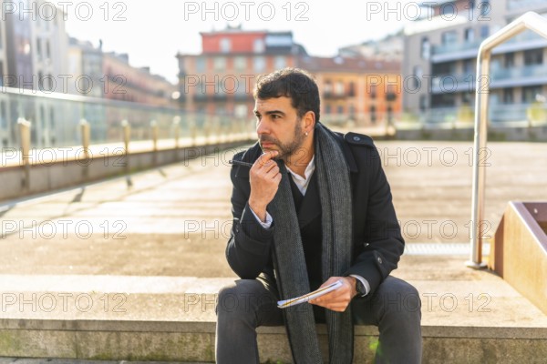 Casual businessman sitting pensive writing notes on notebook outdoors in the city in a sunny day of winter