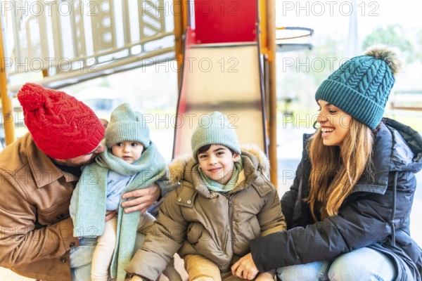 Smiling family with two children wearing winter clothes, hats and scarves enjoying time together on a playground slide