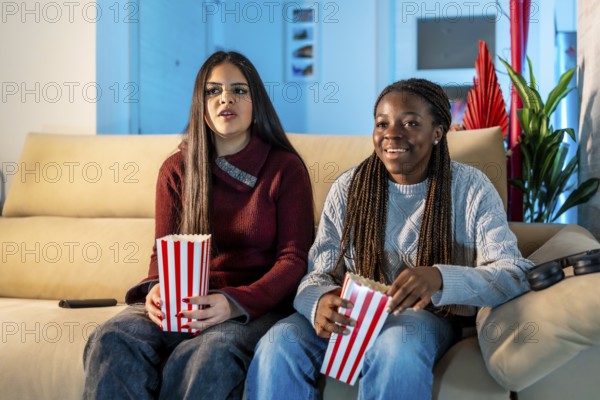Two teenage girls enjoying a movie night at home, sitting on a comfortable sofa, eating popcorn from striped boxes, and having a great time together