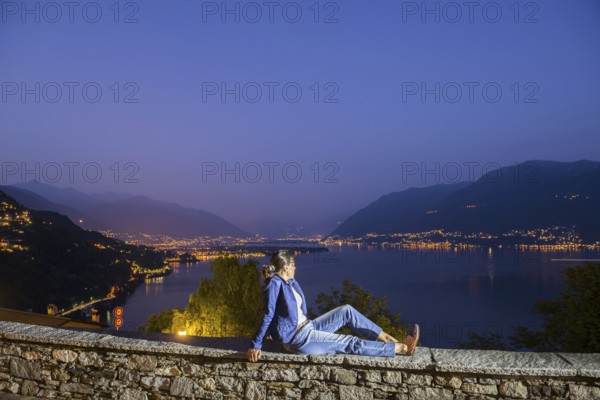 Woman Sitting on a Wall with Panoramic View over Lake Maggiore and Mountain in Dusk in Ronco sopra Ascona, Ticino, Switzerland, Europe
